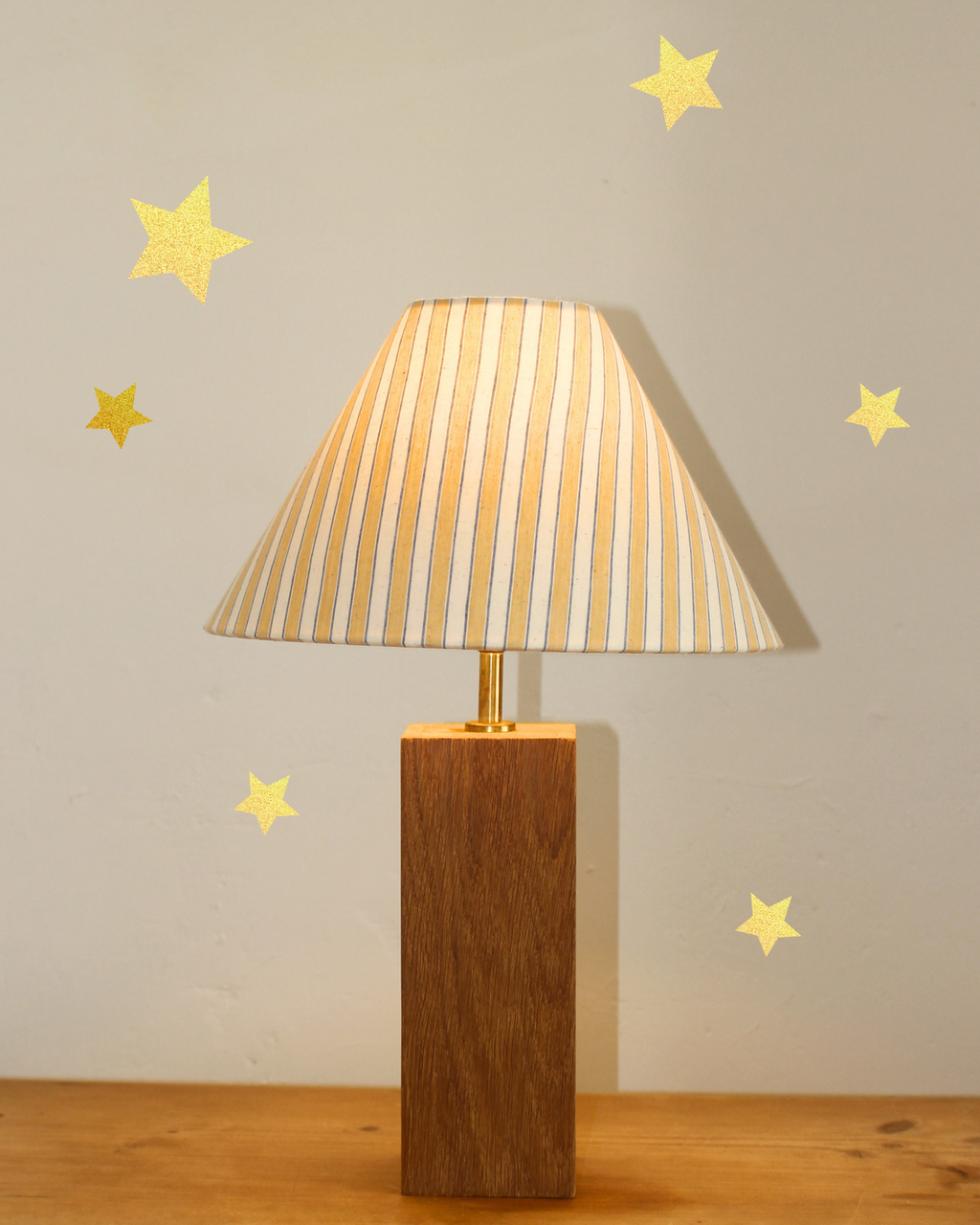 A table lamp with a tapered beige and white striped fabric shade and a rectangular brown wooden base, shown on a wooden surface against a light gray wall with gold star decals.